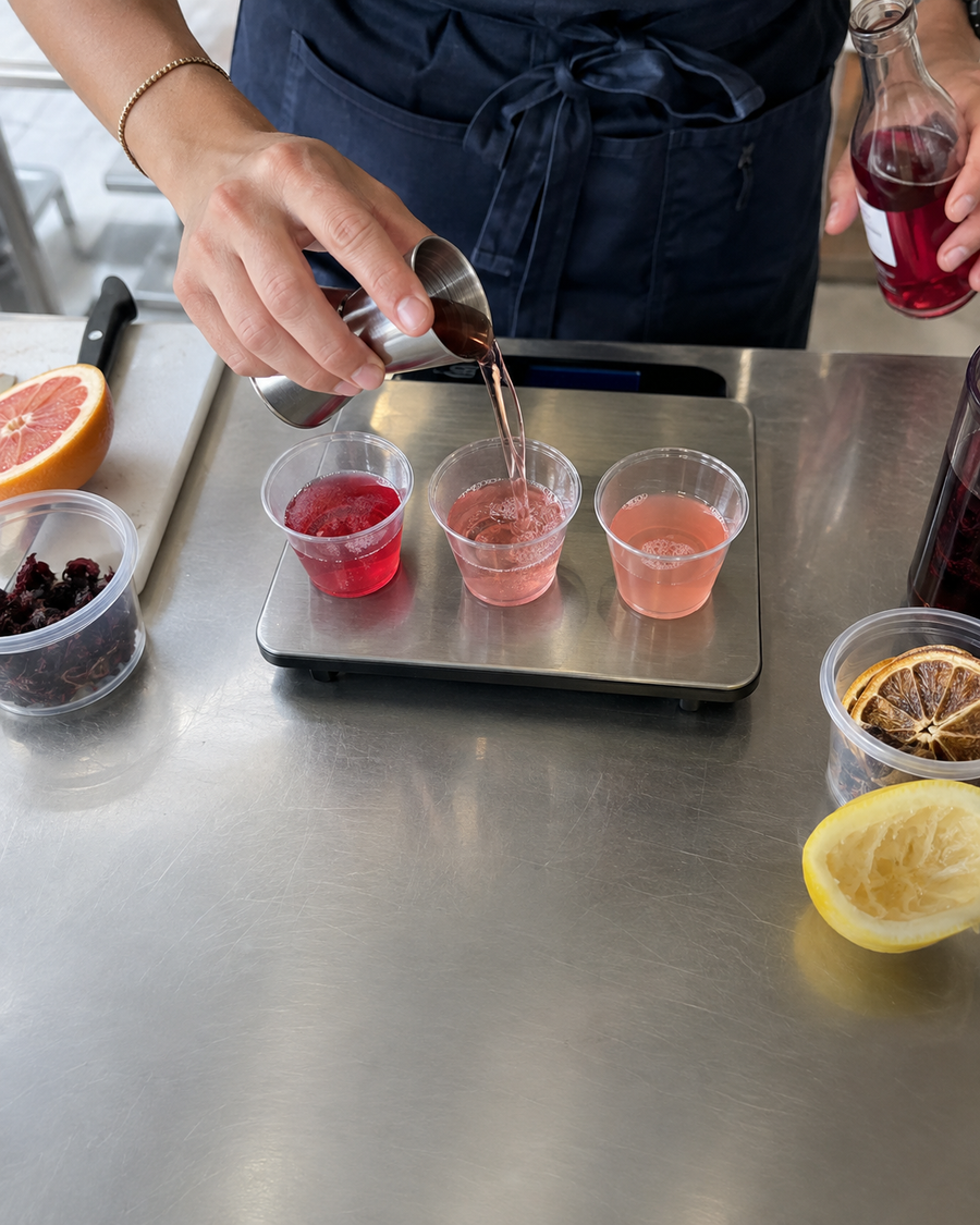Bartender pouring a red mocktail into tasting cups beside fresh citrus and garnishes